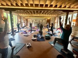 A group of people practice yoga in a spacious, rustic room with wooden beams, brick walls, and natural light. They are all in a lunge pose with arms raised.