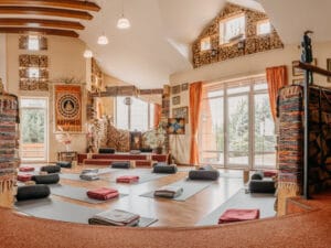 Spacious yoga studio with mats, bolsters, and folded blankets arranged on the floor, natural light from large windows, and stacked firewood decorating the walls.