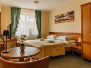 A neatly made bed with folded towels in a hotel room featuring wooden furniture, a round table with chairs, and a window with green curtains.