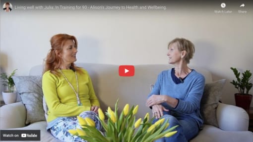 Two women sit on a beige couch having a conversation; a vase of yellow tulips is on a table in the foreground.