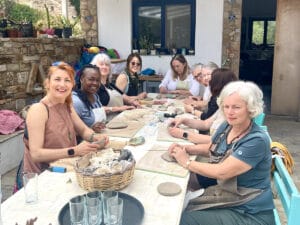 A group of women sit around a table outdoors, working on clay crafts and smiling, with art supplies and finished pieces on the table.