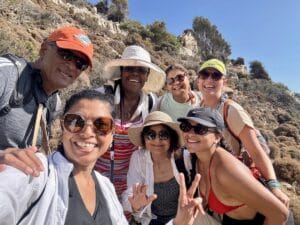A group of seven people pose for a selfie outdoors on a sunny day, wearing sunglasses, hats, and light clothing, with rocky terrain and shrubs in the background.