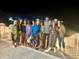 A group of eleven adults pose together outdoors at night on a terrace with empty chairs and tables, with distant lights visible in the background.
