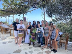 A group of thirteen people pose together outside under a vine-covered pergola next to a row of empty wooden tables and chairs on a sunny day.