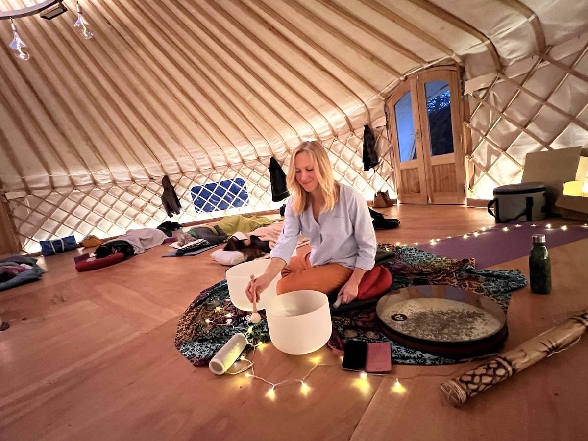 A woman sits on the floor of a softly lit yurt, surrounded by musical instruments, cushions, and string lights, appearing to prepare for a sound meditation session.