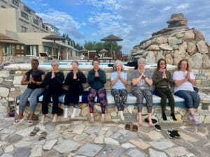 Eight women sit in a row on an outdoor stone bench, eyes closed and hands in a prayer position, with a rocky backdrop and buildings in the background.
