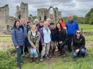 A group of people poses in front of historic stone ruins on a cloudy day, standing and kneeling on grass near a wooden fence.