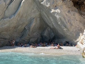 People relax and sunbathe on a small pebble beach at the base of large rocky cliffs, with turquoise water in the foreground.