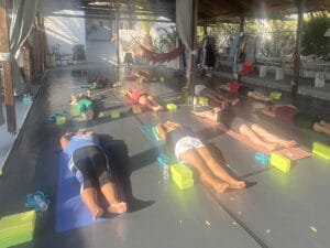 A group of people lying on yoga mats in a spacious, open-air studio, each with yoga blocks and straps beside them, sunlight streaming in from the side.