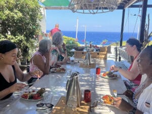 A group of people sit around a table outdoors, eating a meal together with a view of the ocean in the background on a sunny day.