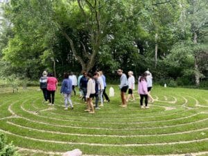 A group of people walk barefoot through a circular grass labyrinth surrounded by trees in an outdoor setting.