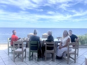 Five women sit at a table on a tiled terrace overlooking the sea, with blue sky and scattered clouds in the background.