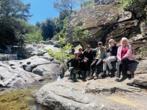 Six people sit together on large rocks beside a clear stream and greenery, enjoying a sunny day outdoors.