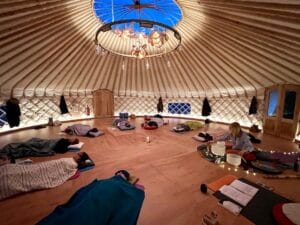 People lie on mats under blankets in a circular wooden yurt, with a teacher near singing bowls. The room is softly lit with candles and overhead lights.