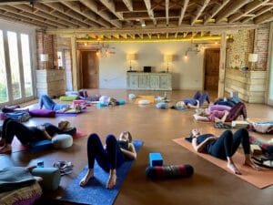 A group of people lie on yoga mats in a spacious, rustic room with exposed beams, practicing relaxation or yoga.