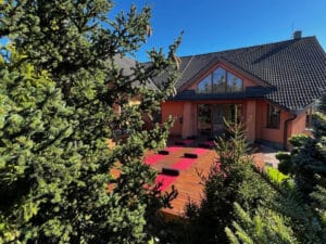 A wooden deck with red cushions is set up outside a house with a tiled roof, surrounded by evergreen trees under a clear blue sky.