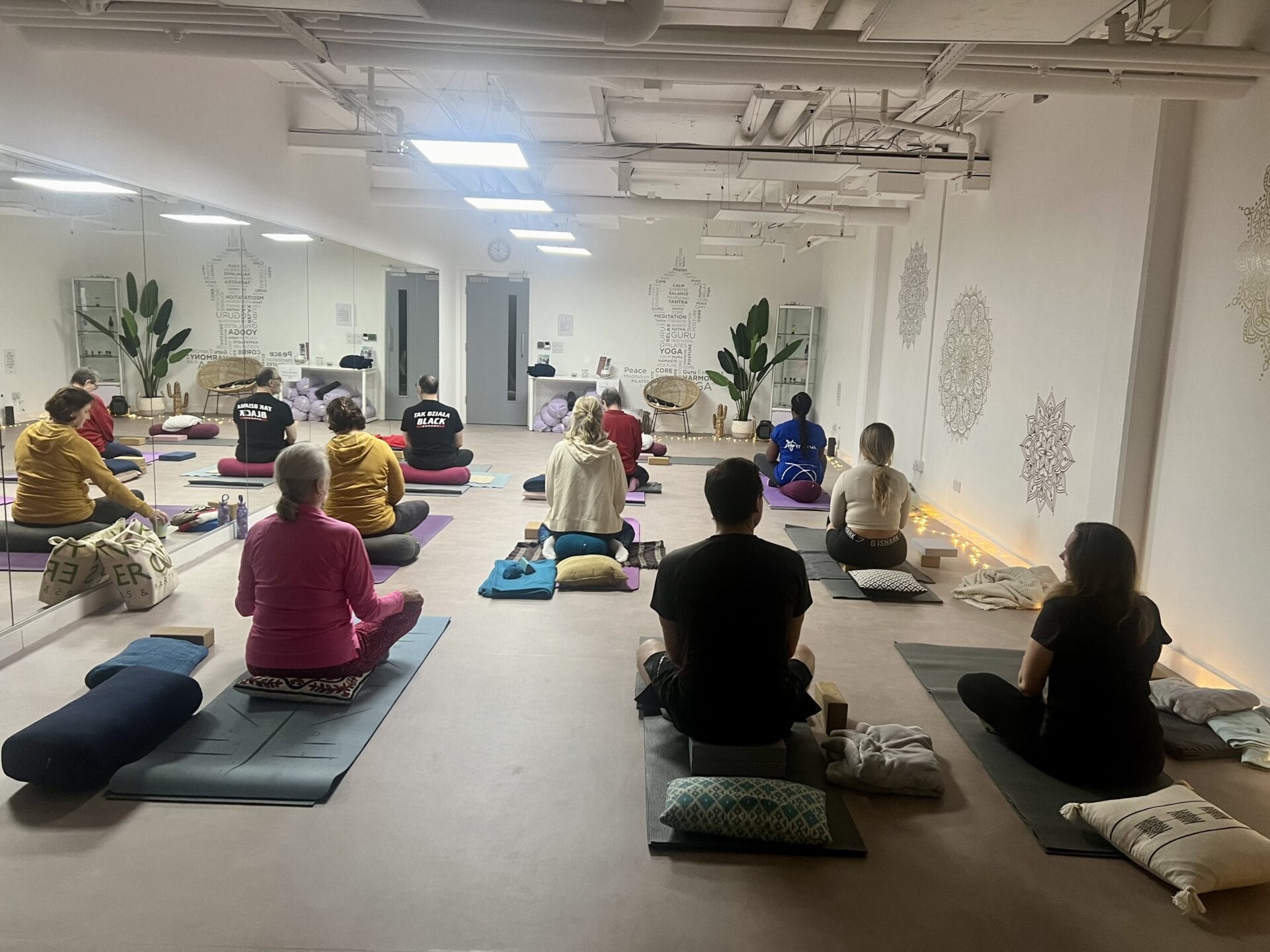 A group of people sit cross-legged on yoga mats in a well-lit studio, facing forward. The room has white walls, plants, and decorative designs.