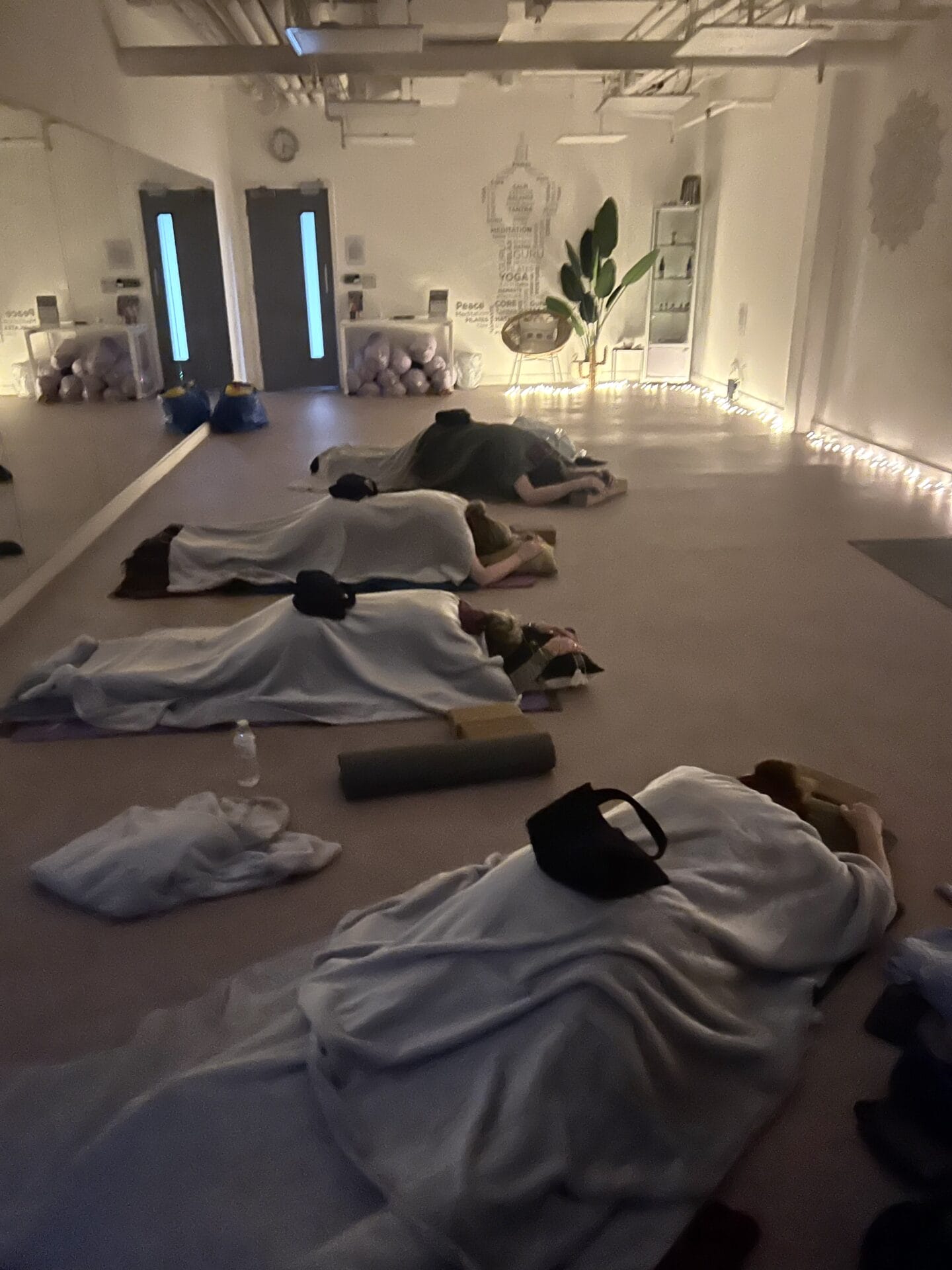 Several people lie on the floor of a dimly lit studio, covered in blankets, with yoga mats and props nearby.