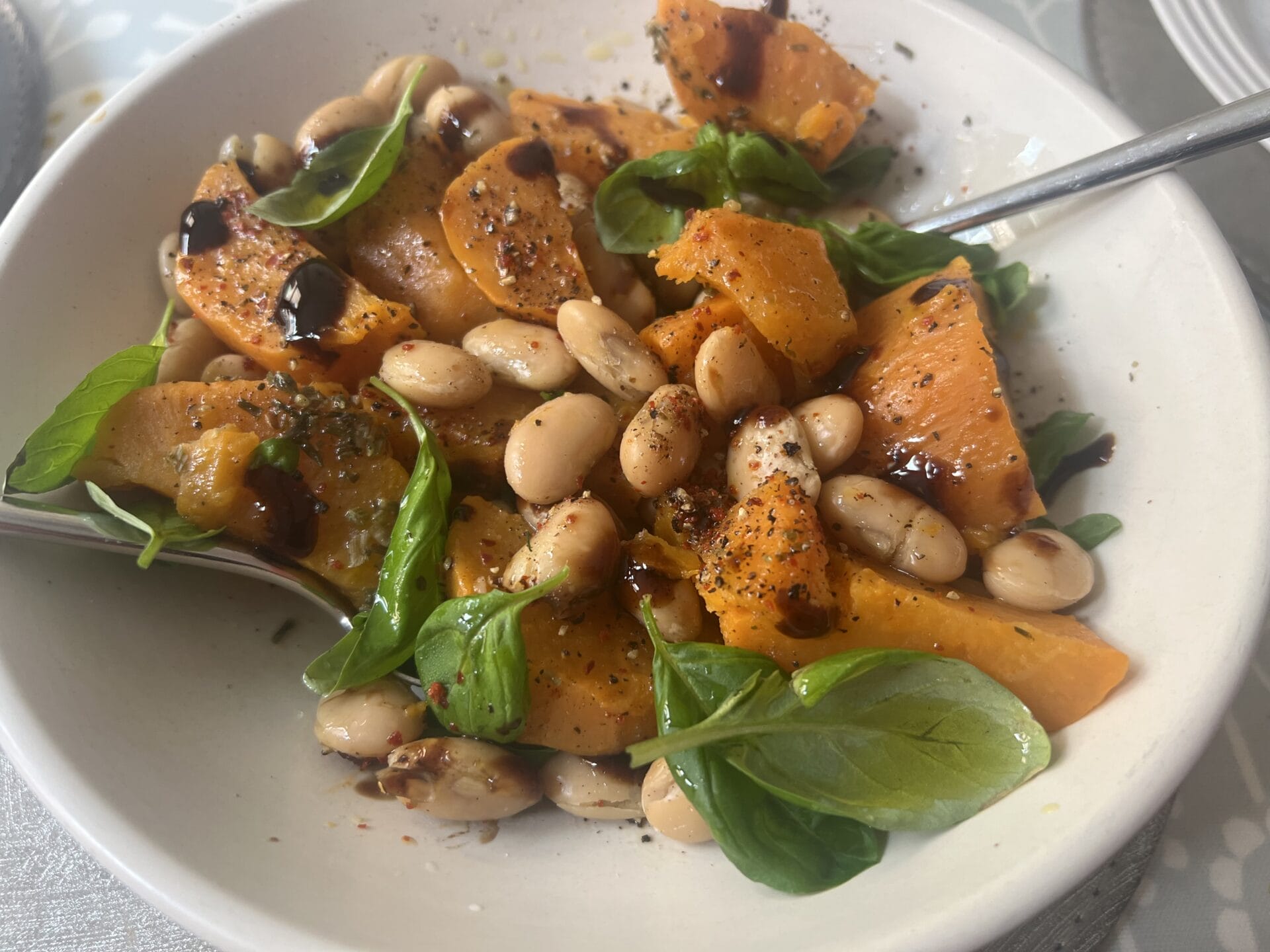 A bowl containing sliced sweet potatoes, white beans, green leaves, and a drizzle of balsamic glaze, with a spoon and fork.
