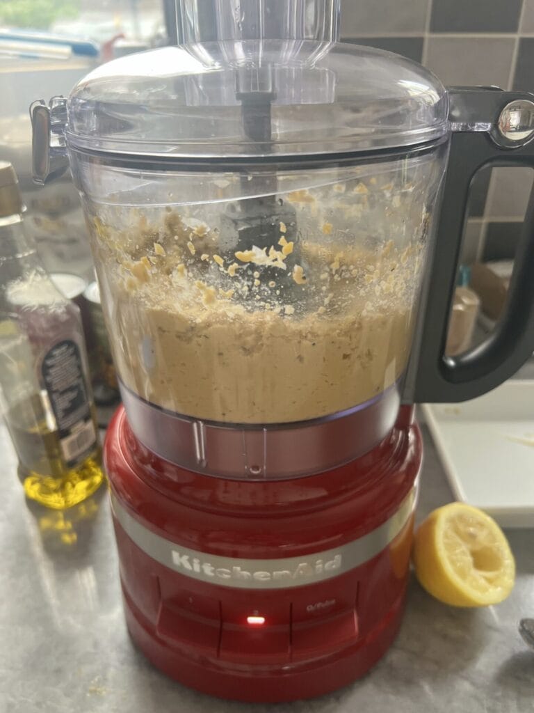 A red KitchenAid food processor blending chickpeas on a kitchen counter, with a bottle of olive oil and a squeezed lemon nearby.