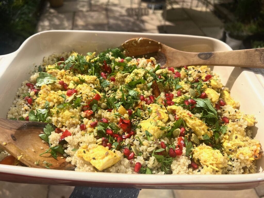 A dish of quinoa salad topped with herbs, pomegranate seeds, and pieces of yellow-seasoned paneer, with wooden serving utensils in the tray.