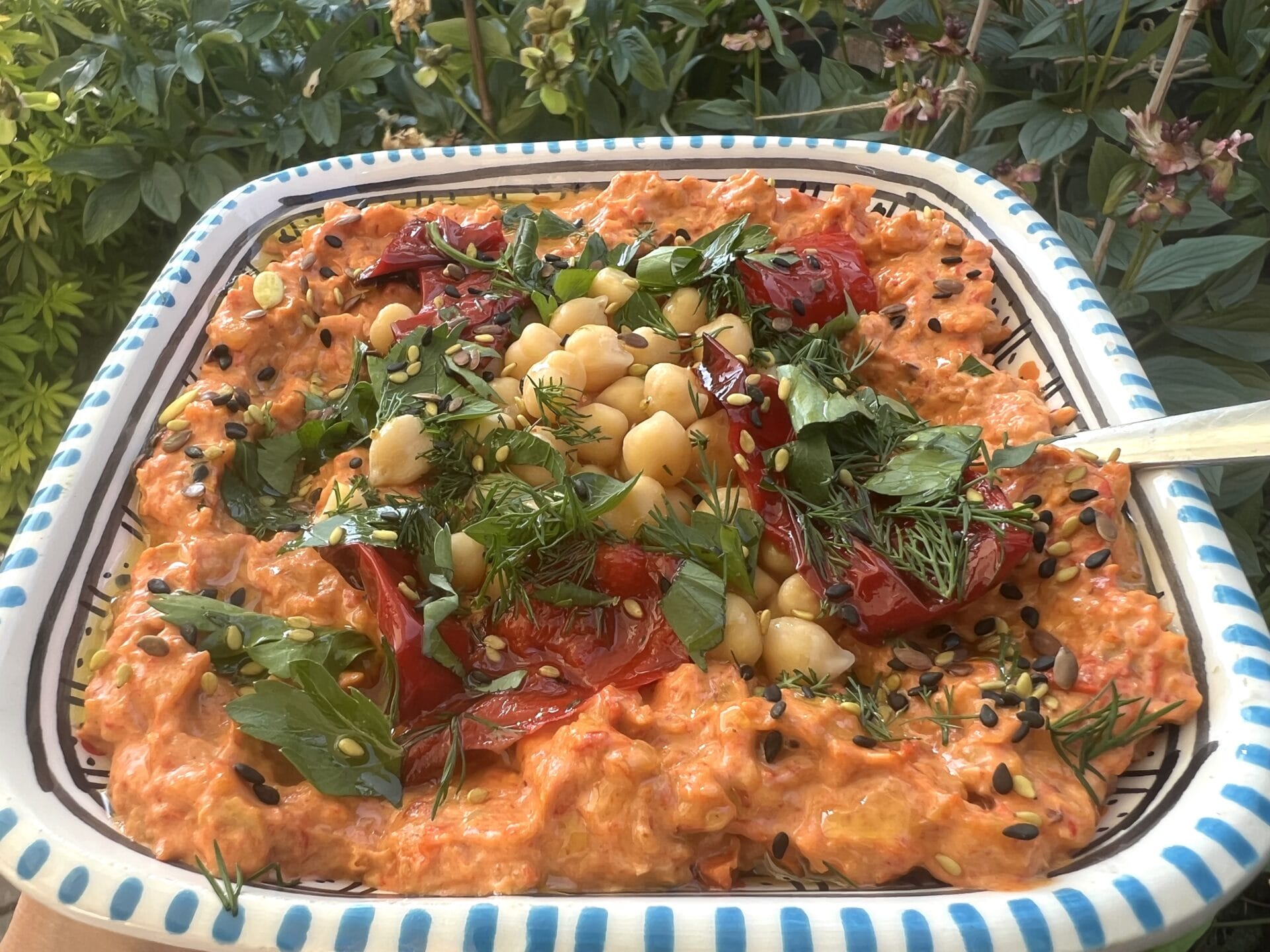 A square dish of creamy red pepper dip topped with chickpeas, herbs, black seeds, and roasted red peppers, set against a background of green foliage.