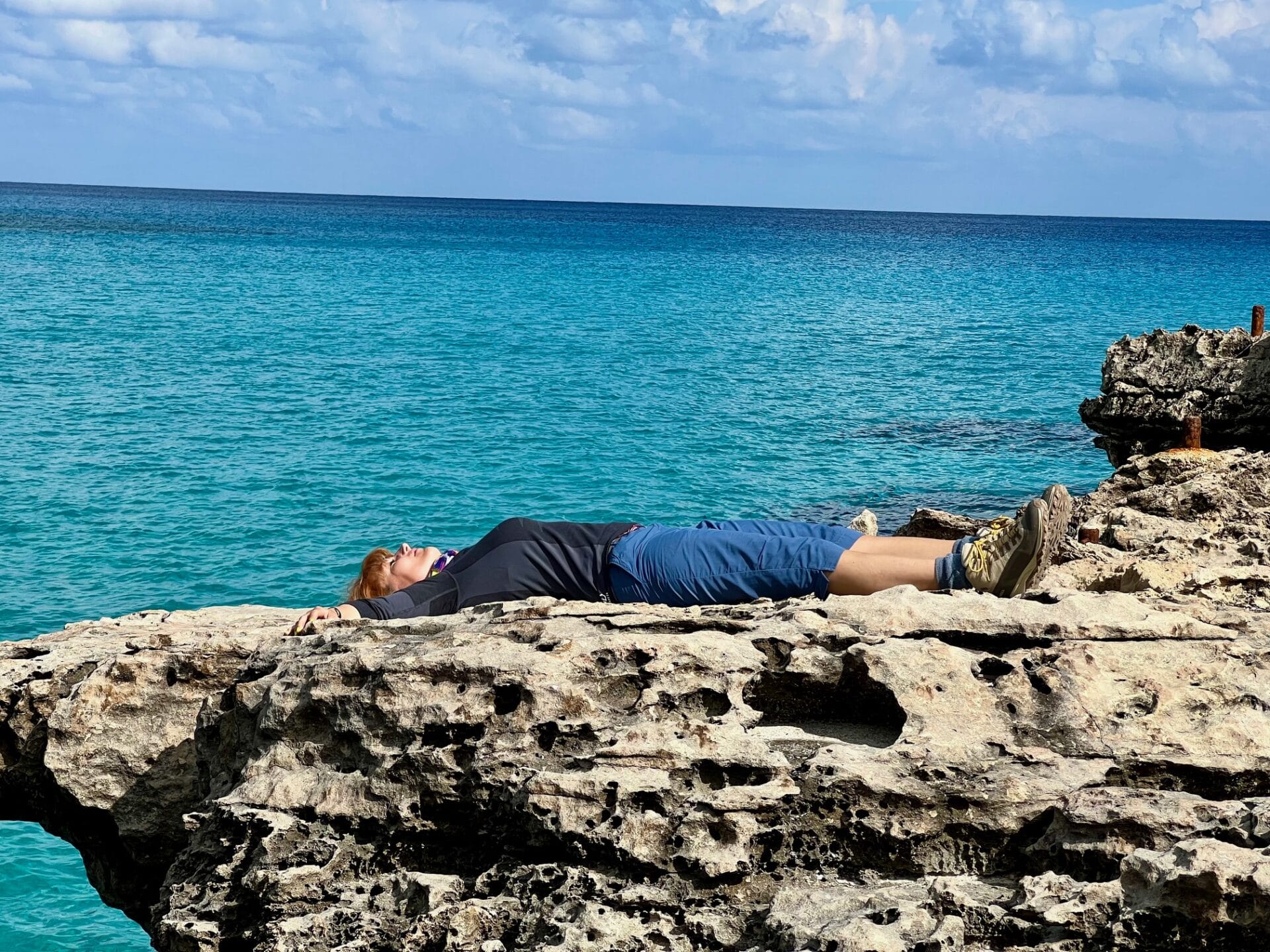 A person lies on their back on a rocky ledge by the turquoise sea under a partly cloudy sky.