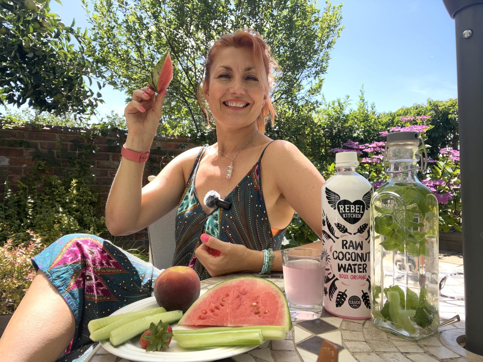 A woman sits outdoors at a table with her 5 hydrating favourites to keep you cool this summer: watermelon, cucumber, a peach, a refreshing drink, raw coconut water, and mint water. She smiles brightly while holding a juicy slice of watermelon.