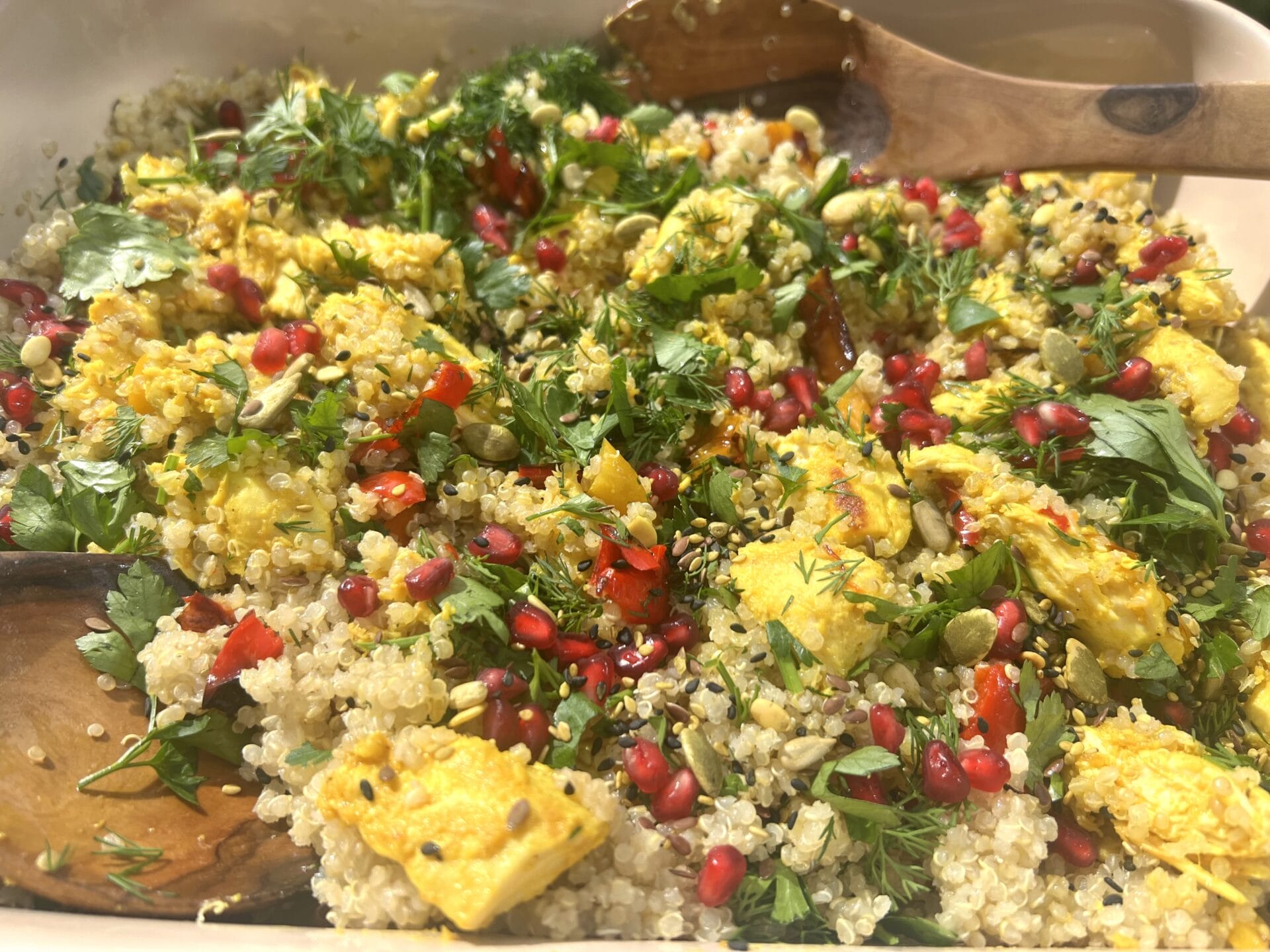 A Glutamine-Rich Gut Healing Dish, this quinoa salad features yellow roasted vegetables, fresh herbs, pomegranate seeds, and mixed seeds, all served in a white tray with wooden utensils.