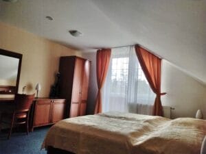 A bedroom with a double bed, wooden furniture, a mirror, a desk with a chair, and a window with orange curtains letting in natural light.