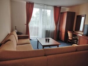 A living room with a beige sectional sofa, wooden coffee table, desk with chair, wardrobe, TV, and large window with sheer and orange curtains.