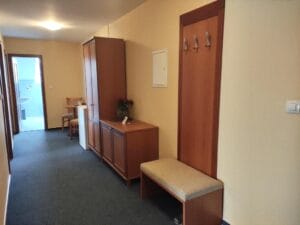 A hallway with blue carpet, wooden furniture including a wardrobe, bench, and coat rack, a small table with chairs, and a doorway leading to another room.