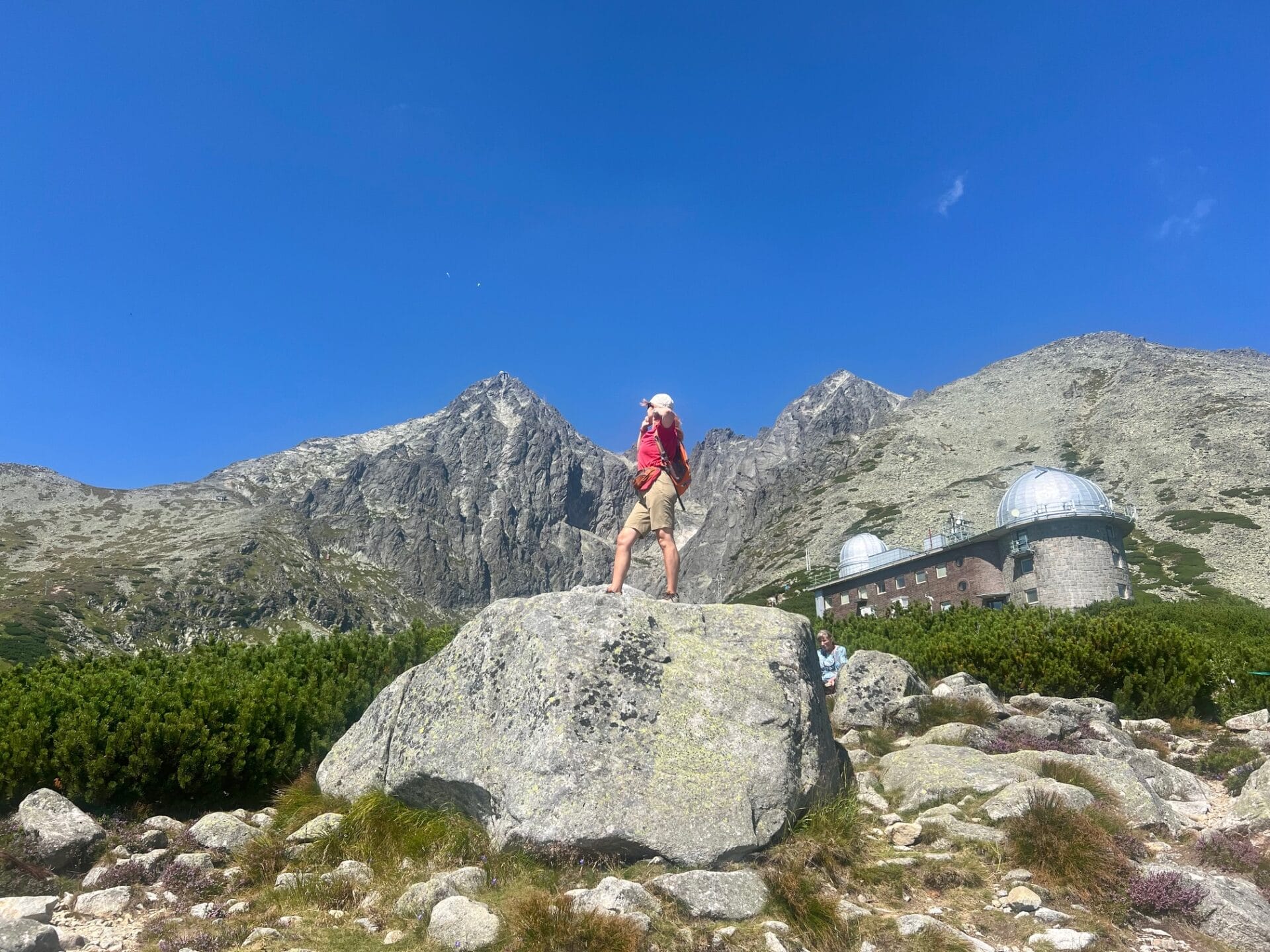 A person stands on a large rock with mountains and an observatory in the background under a clear blue sky.