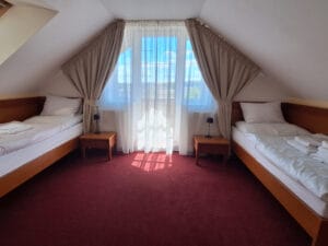 A tidy attic bedroom with two single beds, a bedside table with a lamp between them, red carpet, and a window with sheer curtains letting in sunlight.