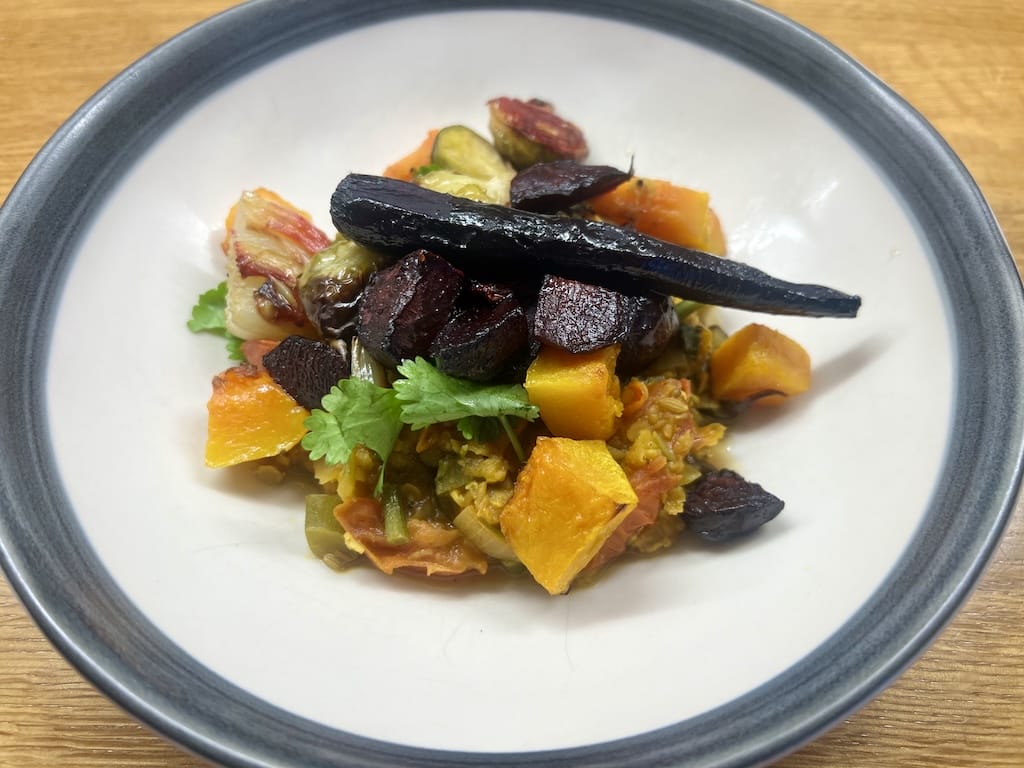 A white bowl contains a mixed vegetable dish with chunks of roasted squash, carrots, herbs, and dark-colored root vegetables on top.