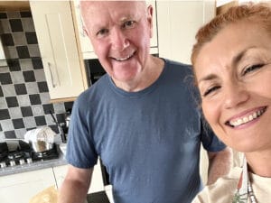 Two people smiling in a kitchen; one man in a blue shirt stands next to a woman taking a selfie. Black-and-white tile backsplash and cooking pot visible in the background.