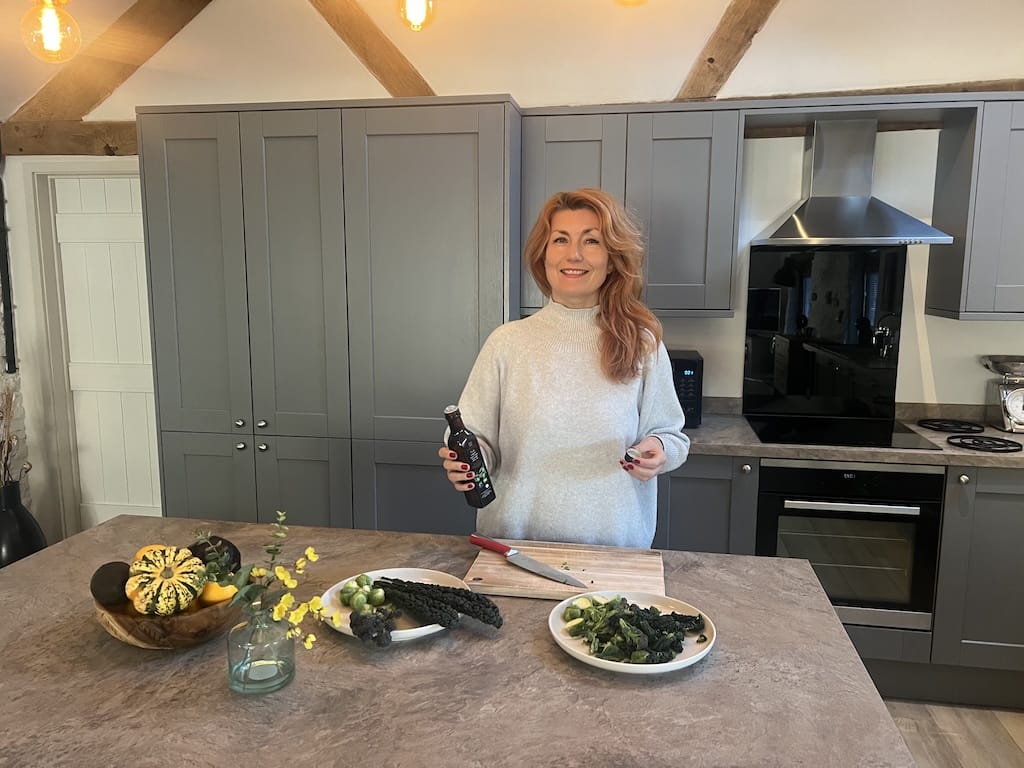 A woman stands in a modern kitchen holding a bottle, with vegetables and a cutting board on the counter in front of her.