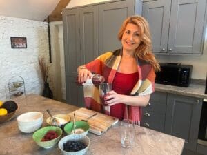 A woman in a red dress and patterned shawl pours juice into a glass in a modern kitchen with bowls of fruit on the counter.