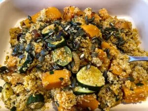 A dish with quinoa, roasted butternut squash, zucchini slices, pumpkin seeds, and a fork on the side.