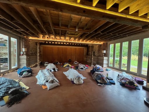 People are lying on mats under blankets in a spacious room with wooden beams, large windows, and natural light, suggesting a group relaxation or meditation session.