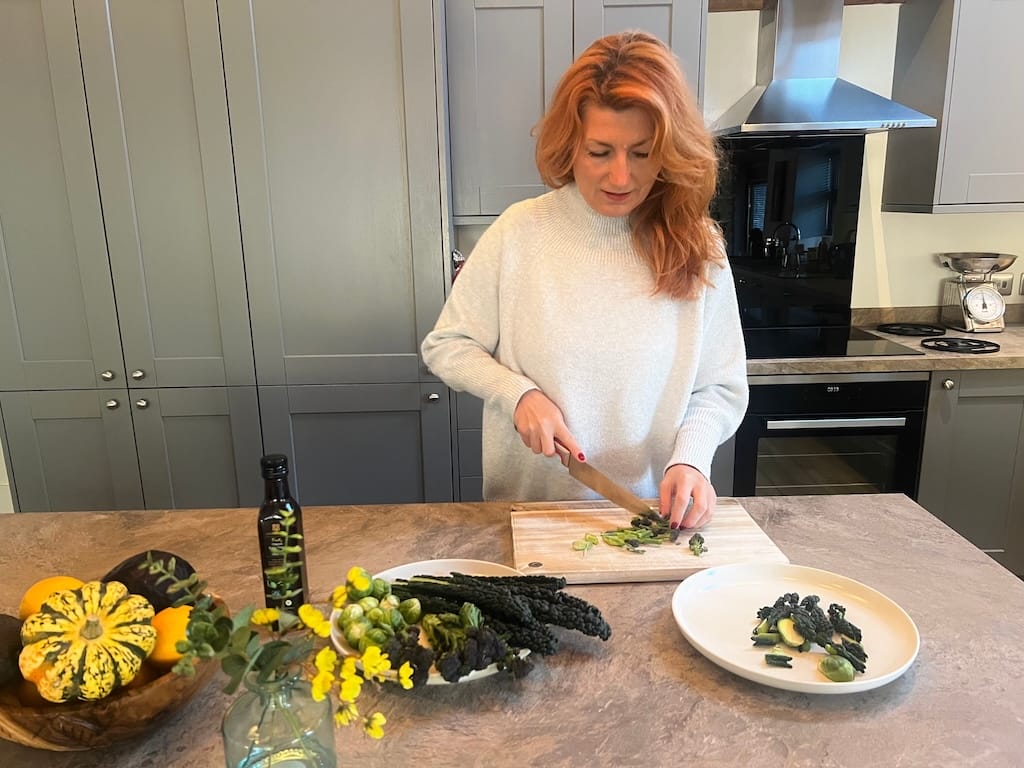 A woman with red hair chops vegetables on a cutting board in a modern kitchen, with assorted produce and olive oil on the counter.