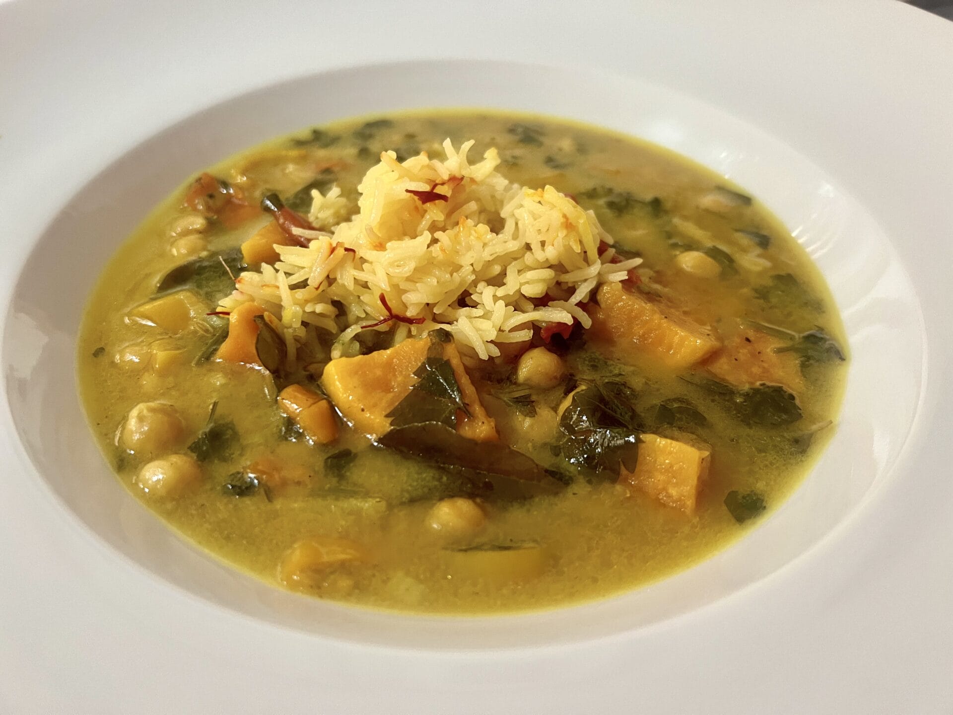 A white plate with a serving of vegetable curry containing chickpeas, spinach, sweet potatoes, and a portion of saffron rice on top.