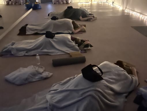 Several people lie on yoga mats under blankets in a dimly lit room, with string lights along the wall and yoga props nearby.