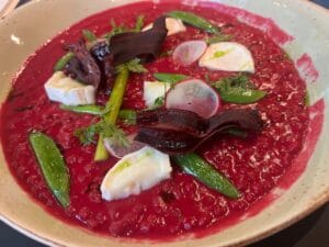 A bowl of beetroot risotto topped with sugar snap peas, sliced radish, pieces of cheese, and garnished with herbs and beet chips.