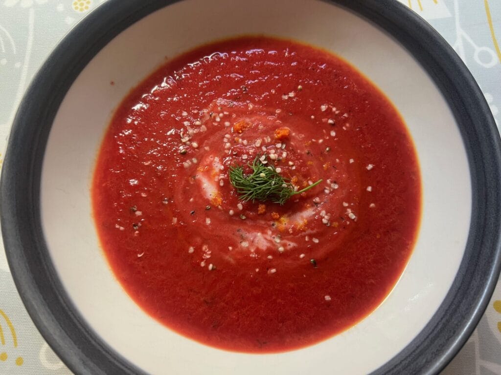 A bowl of red tomato soup garnished with fresh herbs and a sprinkle of grated cheese, served in a white and gray-rimmed bowl.