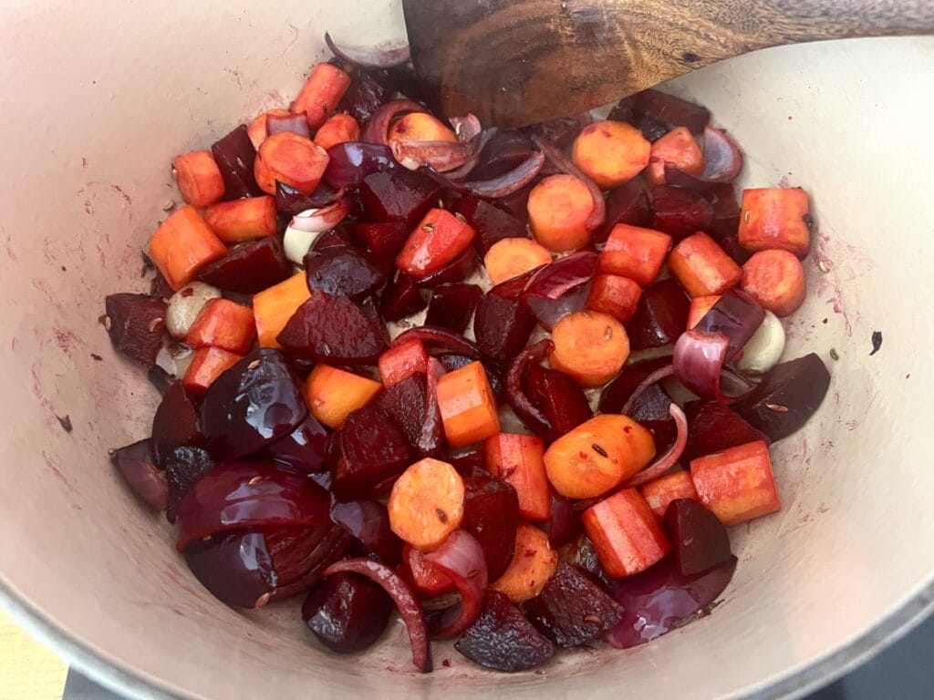 Chopped beets, carrots, red onions, and whole garlic cloves simmer in a pot with a wooden spoon—Goddess Roots Soup, warming and balancing as the season turns.