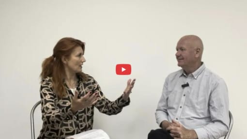 A woman and a man sit facing each other against a plain white background, engaged in conversation. The woman gestures with her hands while the man listens. A play button overlay is visible.