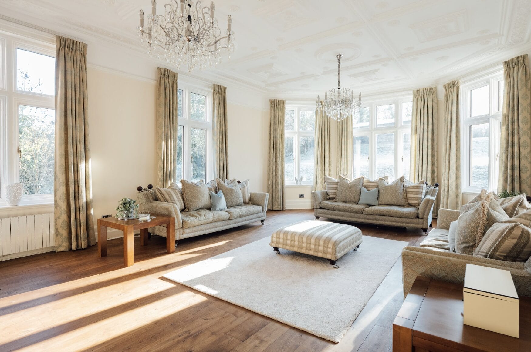 Spacious living room with three sofas, large windows, two chandeliers, beige curtains, wooden floors, and a central ottoman on a white rug.