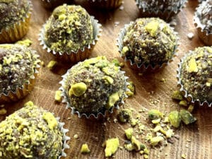 Chocolate truffles coated with crushed pistachios are arranged in gold paper cups on a wooden surface.
