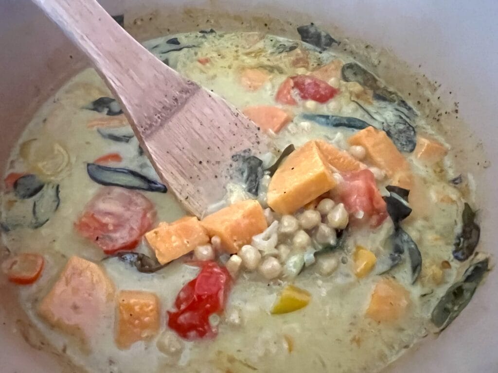 A wooden spoon stirs a creamy Soulful Sweet Potato & Chickpea Curry with tomatoes, greens, and yellow squash simmering together in a pot.